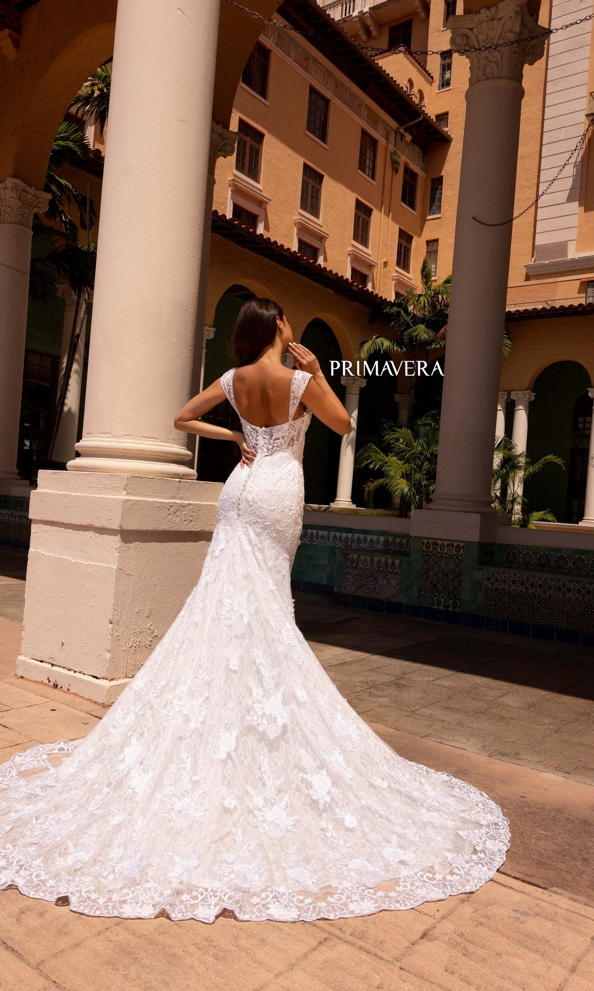 A woman wearing the Long Wedding Dress 11107 by Primavera, featuring delicate cap sleeves in ivory, stands with her back to the camera outside a Mediterranean-style building.